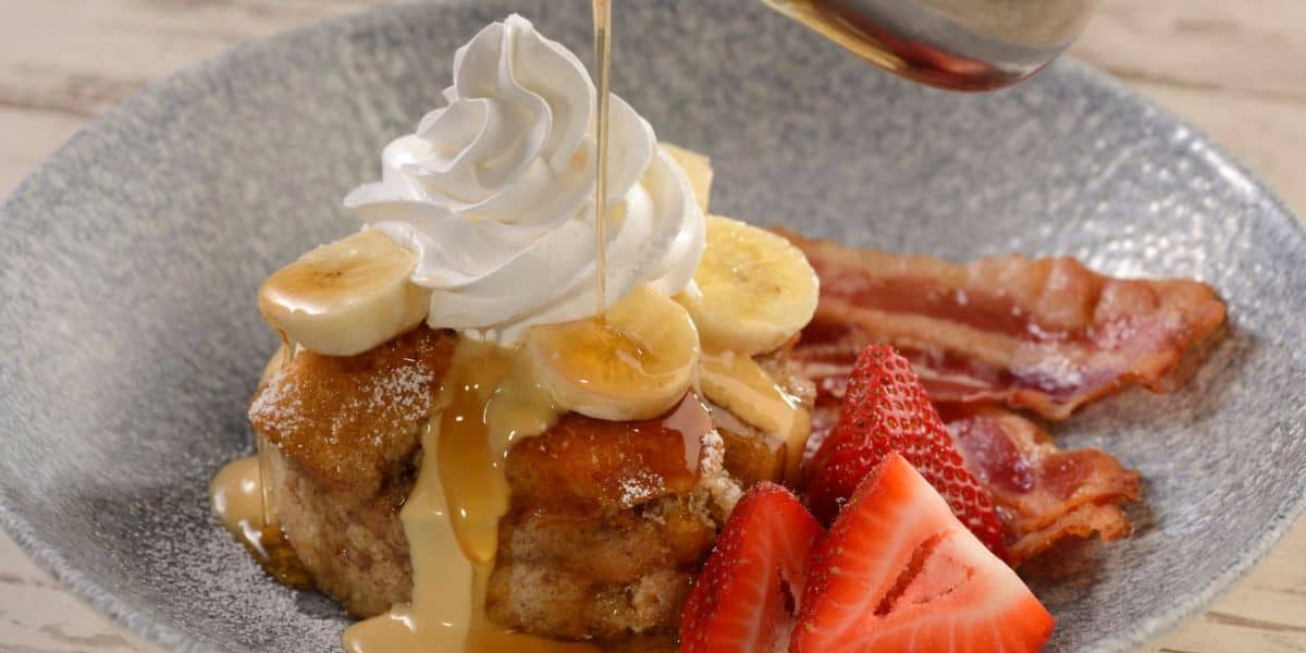 A whimsical brunch plate straight from Walt Disney World, featuring fluffy pancakes topped with banana slices and whipped cream, drizzled with syrup, served alongside fresh strawberries and two crispy strips of bacon—a magical taste of Disney’s breakfast favorites.