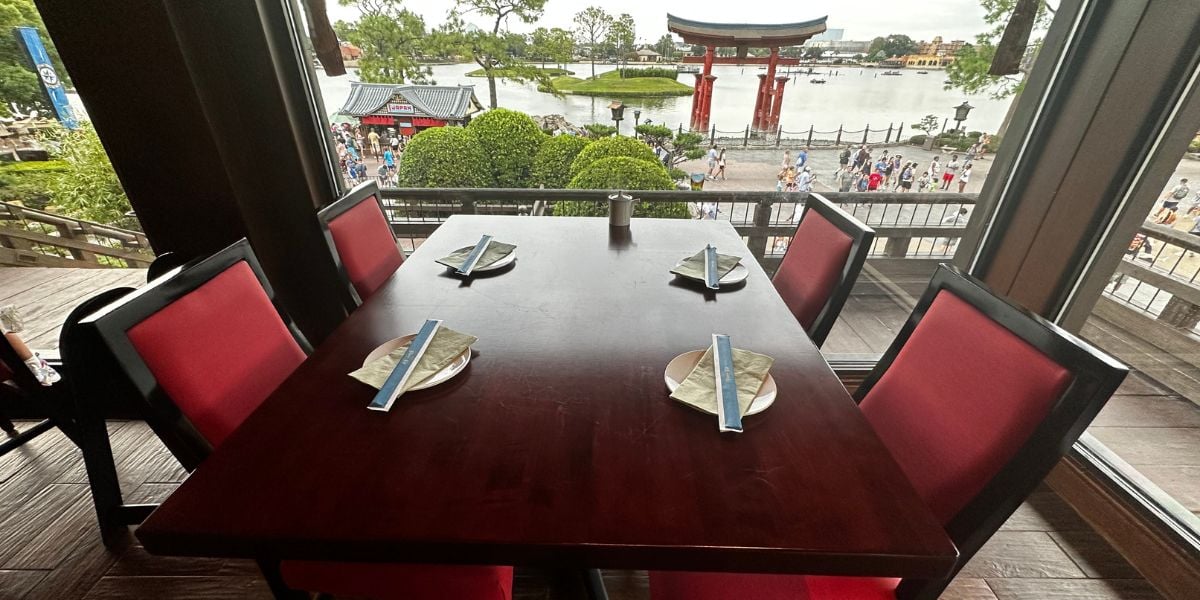 A dining table with four red chairs offers a stunning view of the lake and iconic red torii gate at EPCOT’s Japan Pavilion, creating the perfect atmosphere for guests experiencing the Disney Fireworks Dinner Package with advance reservations.