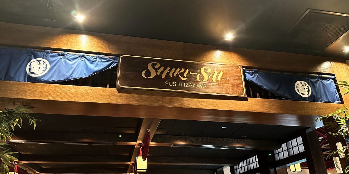 The “SHIKI-SHI SUSHI IZAKAYA” wooden sign welcomes guests above elegant blue noren curtains inside the EPCOT Japan Pavilion, inviting visitors to experience a memorable Disney Fireworks Dinner Package.