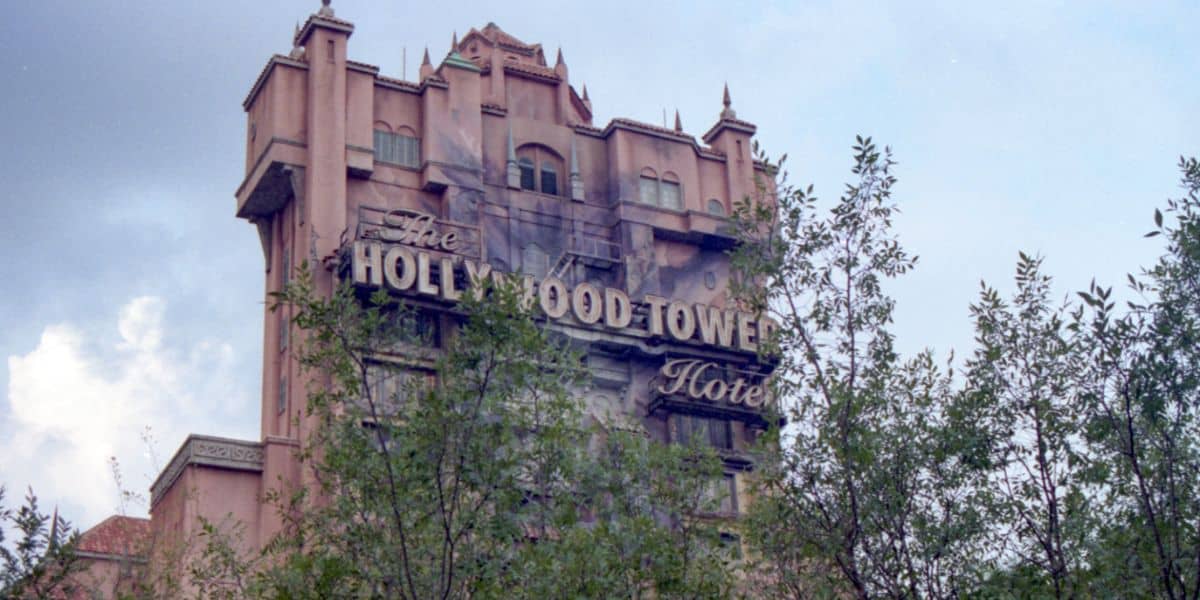 The iconic Hollywood Tower Hotel from Disney’s Tower of Terror attraction looms in a weathered, eerie state, its facade partially hidden by lush theme park foliage beneath a moody, overcast sky.