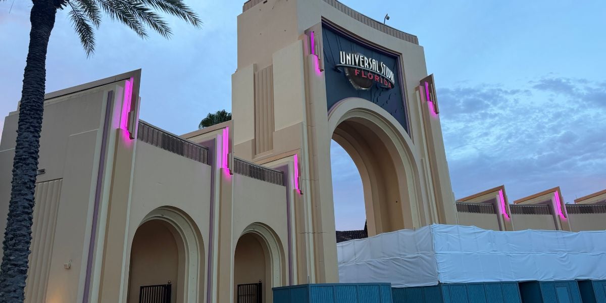 The iconic Universal Studios Florida archway glowing with pink lights at sunset, framed by towering palm trees and temporary construction barriers.