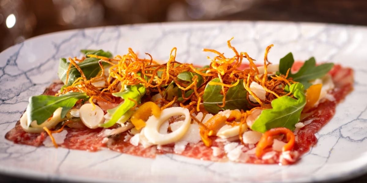 Step into the magic of Disney’s Yacht Club Resort and treat yourself to a show-stopping dish at Yachtsman Steakhouse: thinly sliced beef carpaccio layered with crisp greens, fried carrots, colorful peppers, onions, and cheese, artfully arranged on a white plate. This savory creation is a star among Disney dining experiences—perfect for foodies exploring every epic bite in the parks.