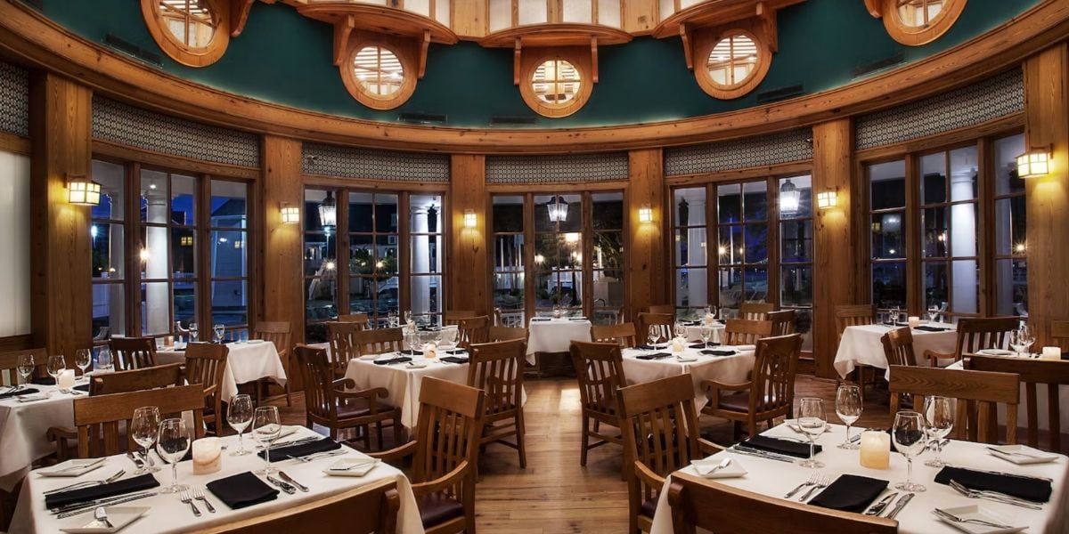 Step into the round dining room at Yachtsman Steakhouse in Disney’s Yacht Club, where wooden chairs, crisp white tablecloths, and grand windows set the stage for your next magical meal. Warm lighting enhances the inviting atmosphere, perfect for theme park guests to unwind and savor inventive new dishes in a refined yet relaxed setting after a day at Walt Disney World Resort.