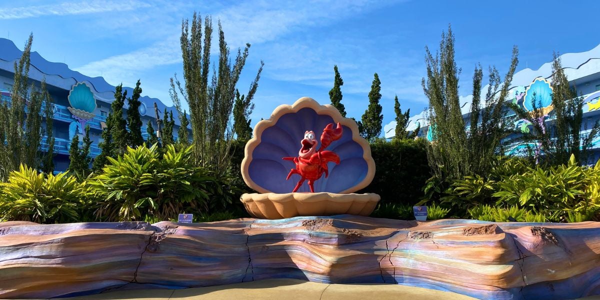 Sebastian the red crab stands inside a giant seashell, surrounded by lush plants and vibrant rocks, creating a lively scene at Disney World’s Art of Animation Resort.