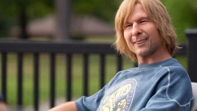Chad Powers flashes a cheerful smile while relaxing outdoors on a chair, sporting his signature blue graphic sweatshirt at the park.