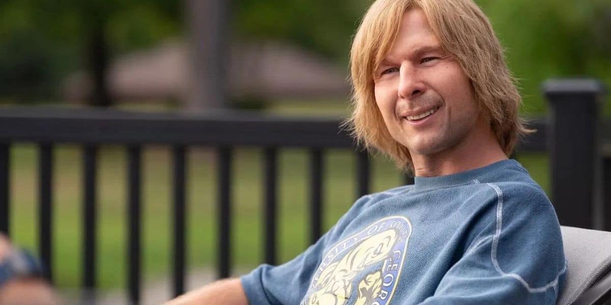 Chad Powers flashes a cheerful smile while relaxing outdoors on a chair, sporting his signature blue graphic sweatshirt at the park.