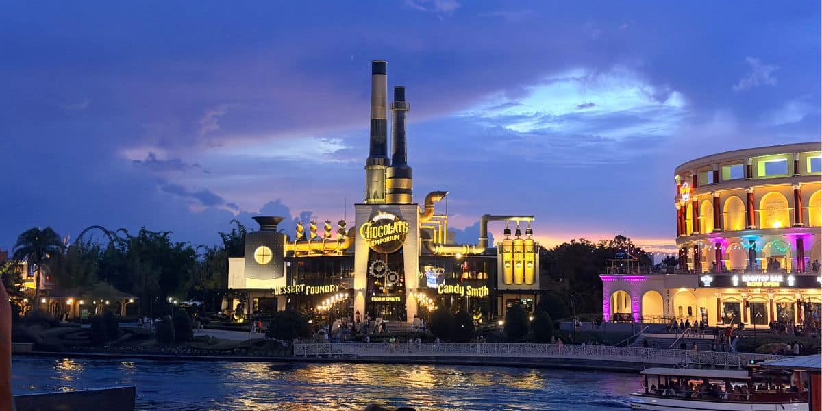 The Chocolate Factory restaurant shines along the riverside at dusk, its inviting lights echoing the vibrant energy of Universal CityWalk. Next door, a brightly illuminated building adds to the excitement of this entertainment hub, where crew members are trained to deliver standout service on par with favorites like Five Guys.