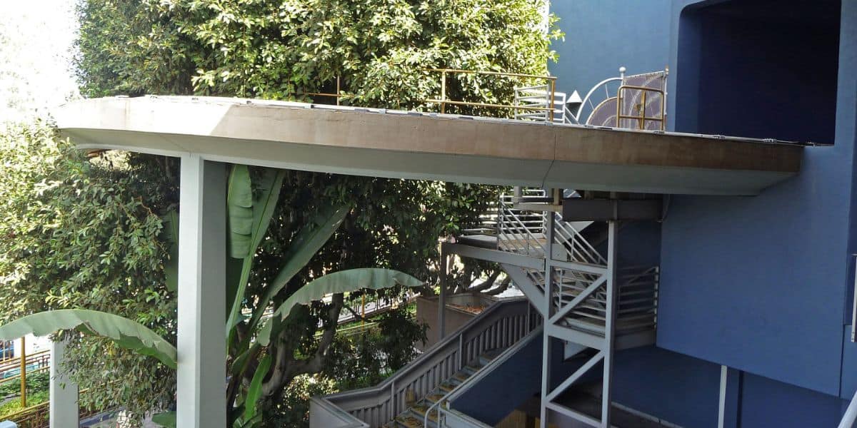 A winding platform with stairs leads out from a vibrant blue pavilion, nestled amid lush themed landscaping and greenery.