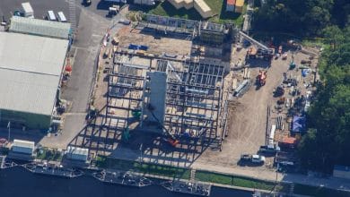 Aerial view of a future theme park under construction, with roller coaster steel framework, colorful ride vehicles, and stacks of materials surrounded by lush trees and nearby attractions.