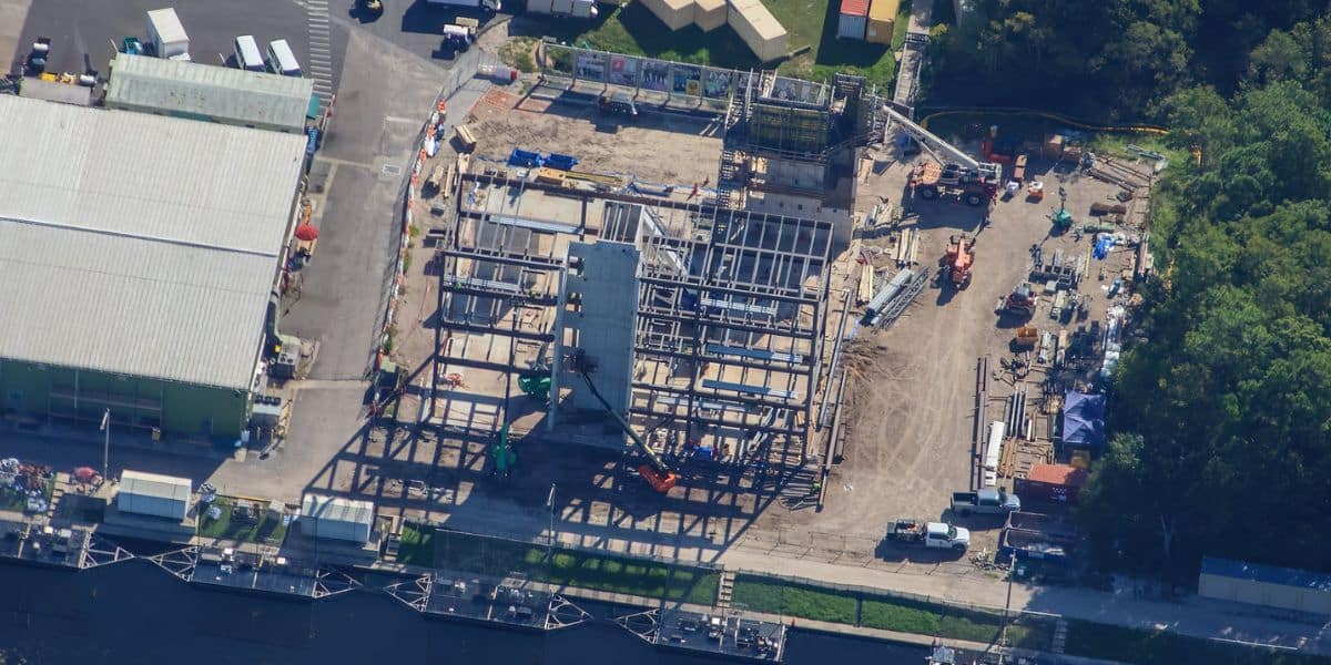 Aerial view of a future theme park under construction, with roller coaster steel framework, colorful ride vehicles, and stacks of materials surrounded by lush trees and nearby attractions.