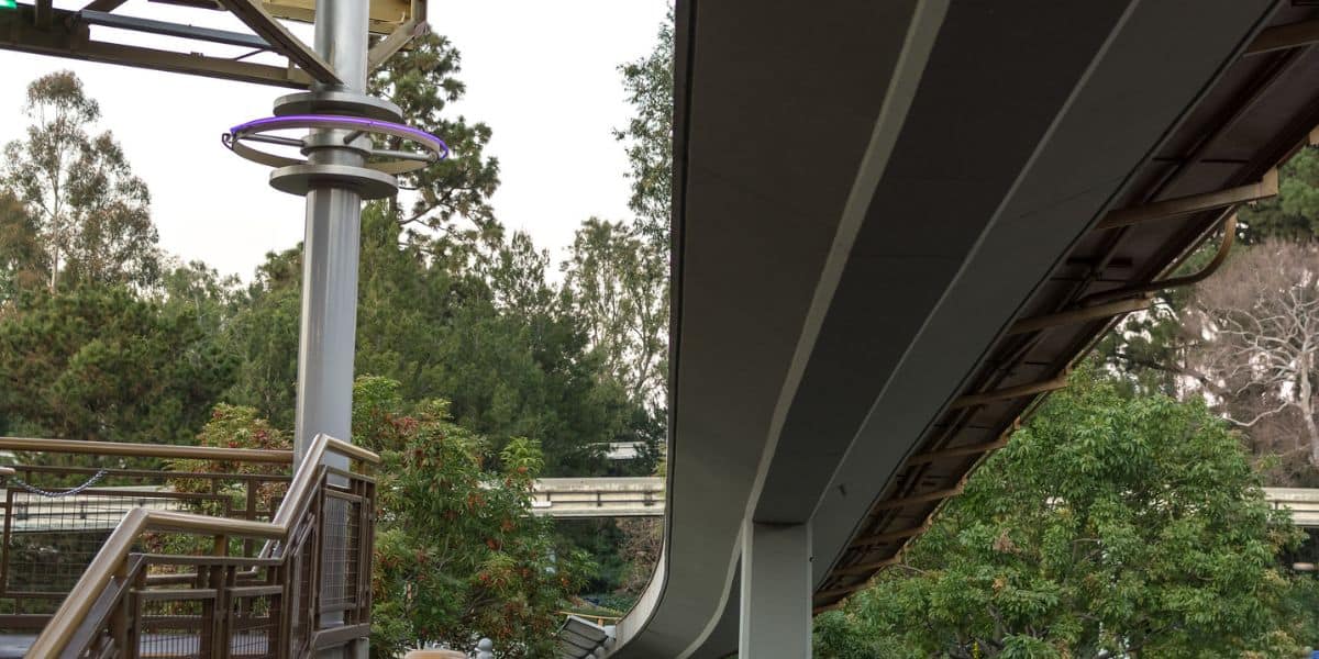 The monorail glides along its elevated track above lush greenery and winding pathways, curving past a glowing purple ring light—an inviting sight for guests exploring the park’s vibrant attractions.