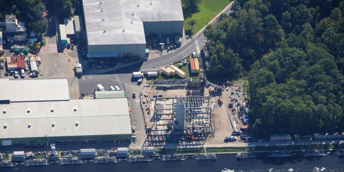 Aerial view of a new roller coaster being constructed with a visible steel framework, located beside large entertainment complexes and bordered by lush wooded areas within a theme park resort.