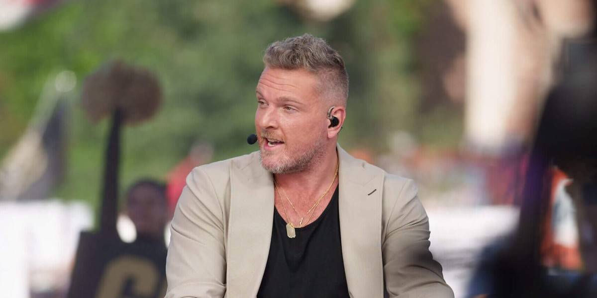 Charismatic host in a beige blazer and black shirt wears a headset while engaging visitors outdoors, vibrant greenery blurred behind him, channeling Pat McAfee’s dynamic style as he entertains the crowd with lively sports banter and playful humor—radiating the high-energy spirit of a Disney Parks show announcer.