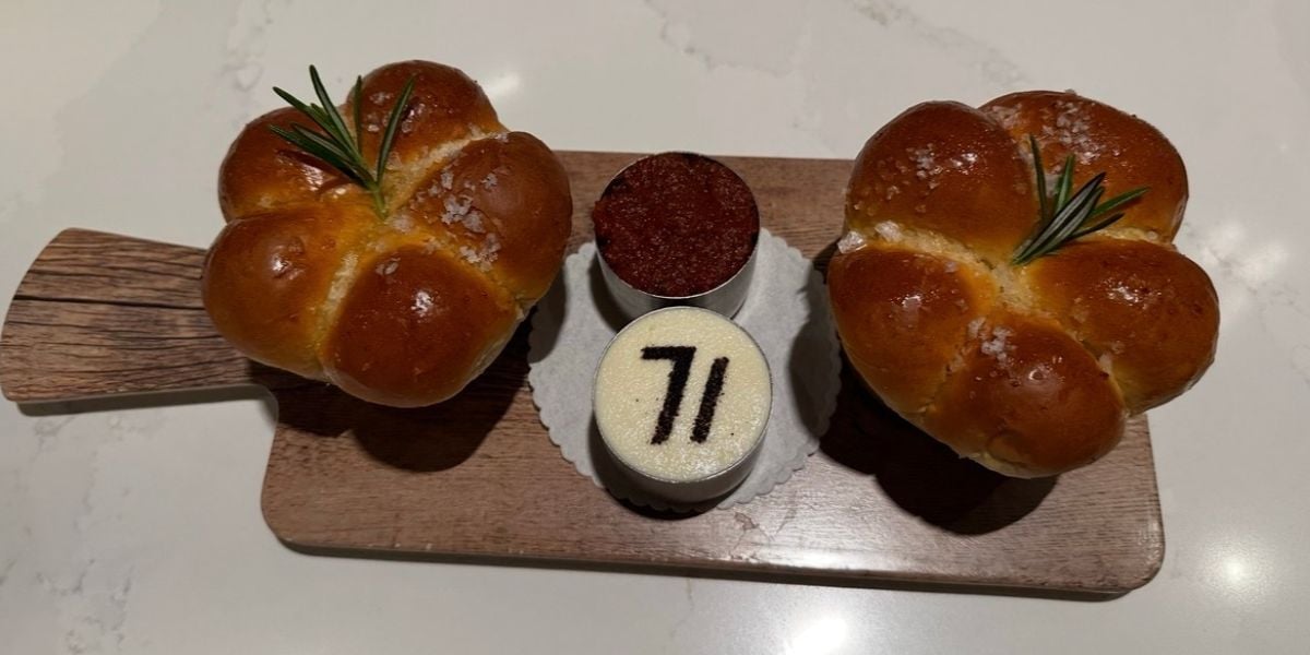 A pair of golden-brown breads sprinkled with rosemary and sea salt sit beside two petite desserts—including one topped with a striking “71”—all beautifully arranged on a wooden board, channeling the upscale culinary flair guests expect at Disney’s Contemporary Resort.