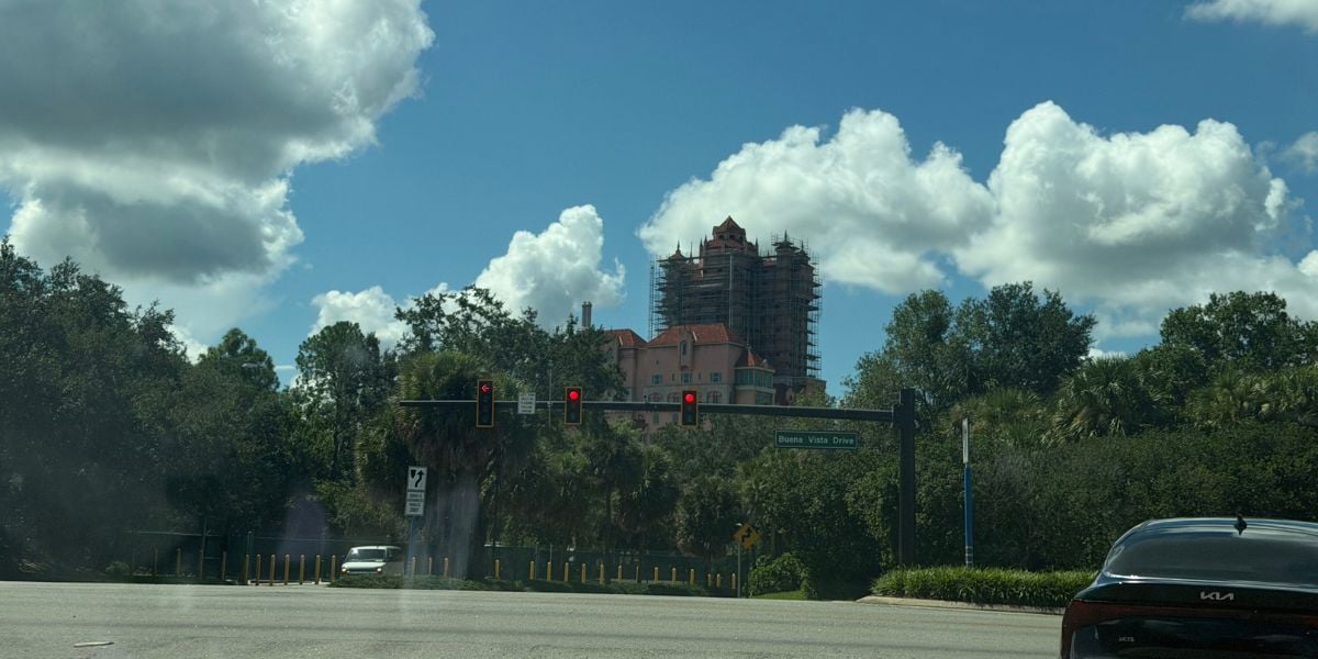 A new attraction rises behind a row of trees, as colorful traffic lights and a Kia pass by at the lively park entrance intersection.