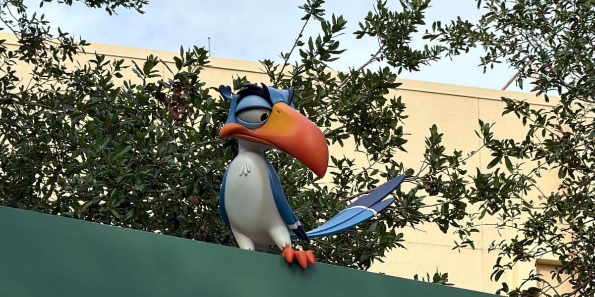 Kazoo, the whimsical bird statue with a bright orange beak from Muppet*Vision 3D, perches playfully on a green ledge at Disney’s Hollywood Studios, surrounded by lush trees and classic park architecture.