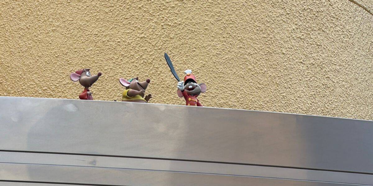 Mickey Mouse, flanked by two classic Disney mouse friends, stands on a ledge at a Hollywood Studios set; Mickey brandishes a shiny silver sword while a textured studio wall creates an authentic movie backdrop.