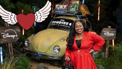 Carla Jeffery, dressed in a vibrant red outfit, beams in front of a vintage car stacked with luggage. Behind her, Disney+ and Disney Channel signs set the scene alongside a playful heart graphic—a delightful tribute to the beloved Disney Channel star that fans will instantly recognize.