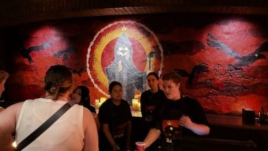 Staff at Jock Lindsey’s Hangar Bar at Disney Springs stand before a large mural of a hooded figure.