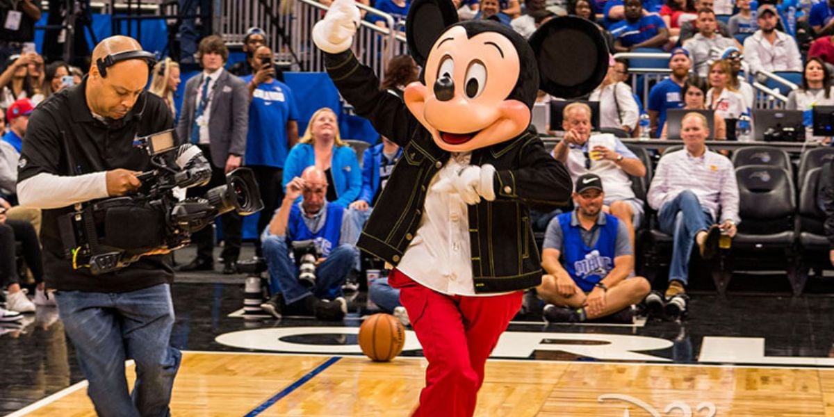 Mickey Mouse waves on the court at a Disney World event held at the Kia Center, while a cameraman captures the moment nearby.